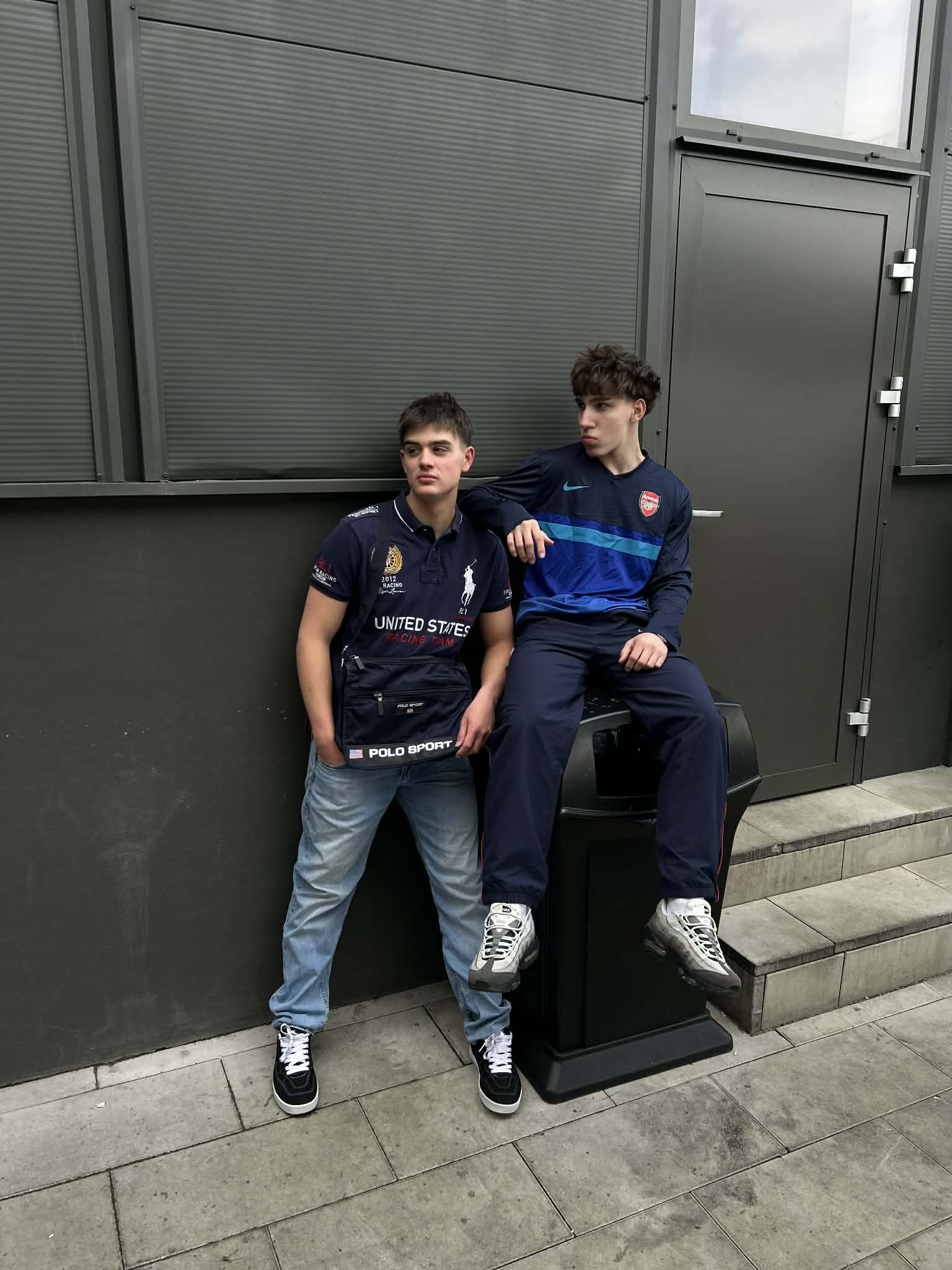 Két fiatal utcai stílusú, vintage ruházatban, szürke háttér előtt pózol.